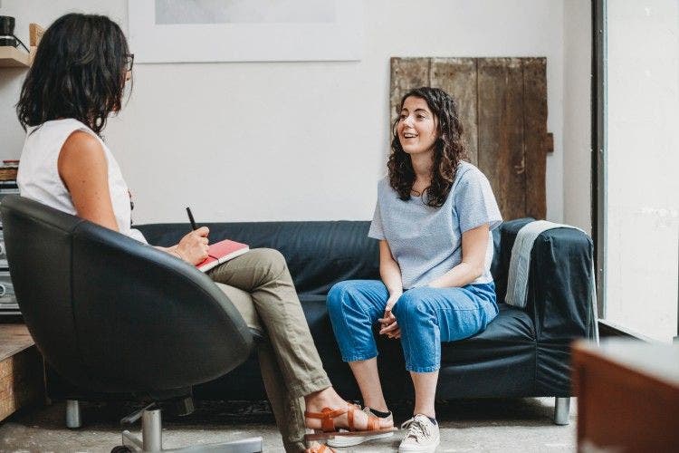 A mental health counselor with a pen and notebook talks with a patient sitting on a couch.
